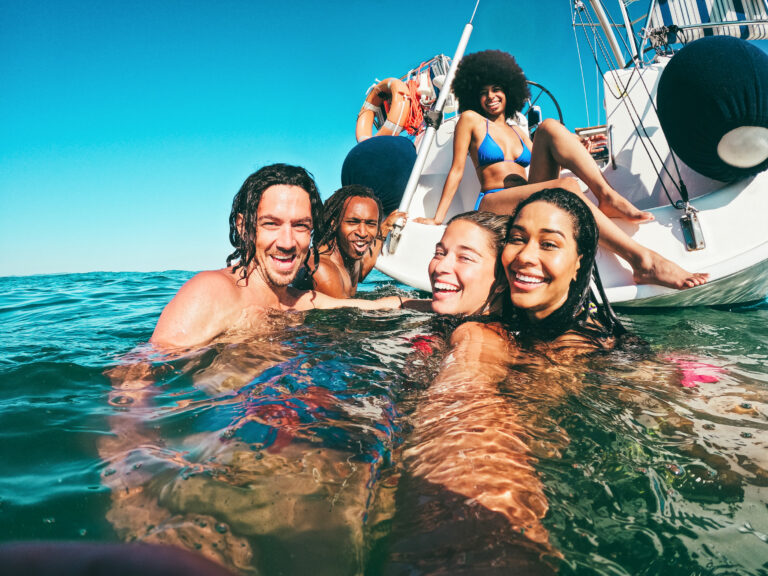 Happy multiracial friends doing selfie swimming in the sea with sail boat in background - Soft focus on center girl face